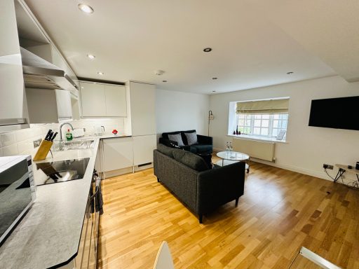 Modern living room with a black sofa, coffee table, and kitchen area, featuring wooden flooring.